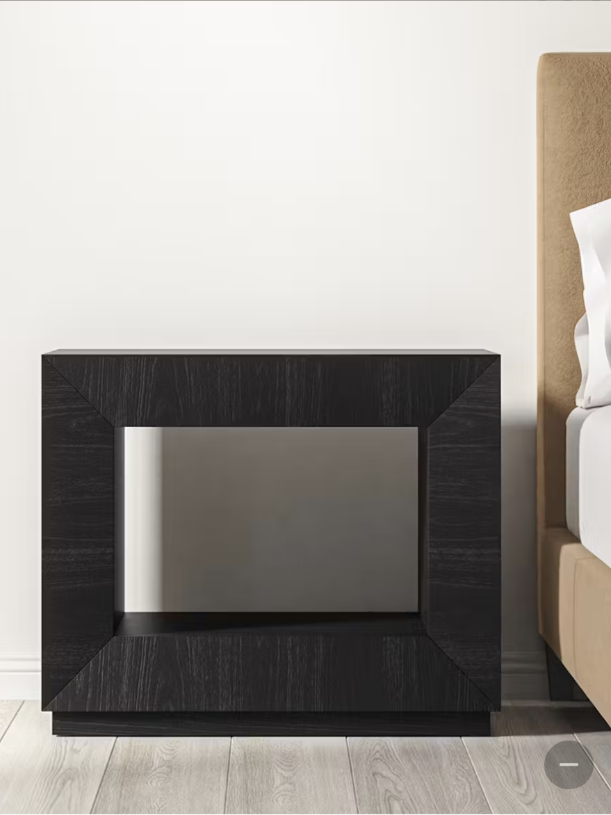 Black geometric nightstand beside a beige upholstered bed with white bedding, on light wooden flooring.
