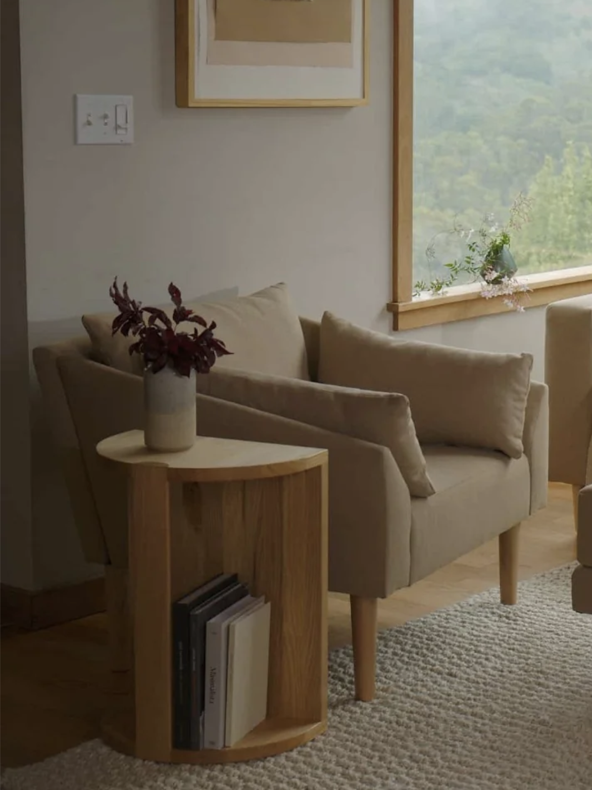 A beige armchair with cushions is next to a small wooden side table holding a vase with dark red flowers and a few books. A window and framed art are visible in the background.