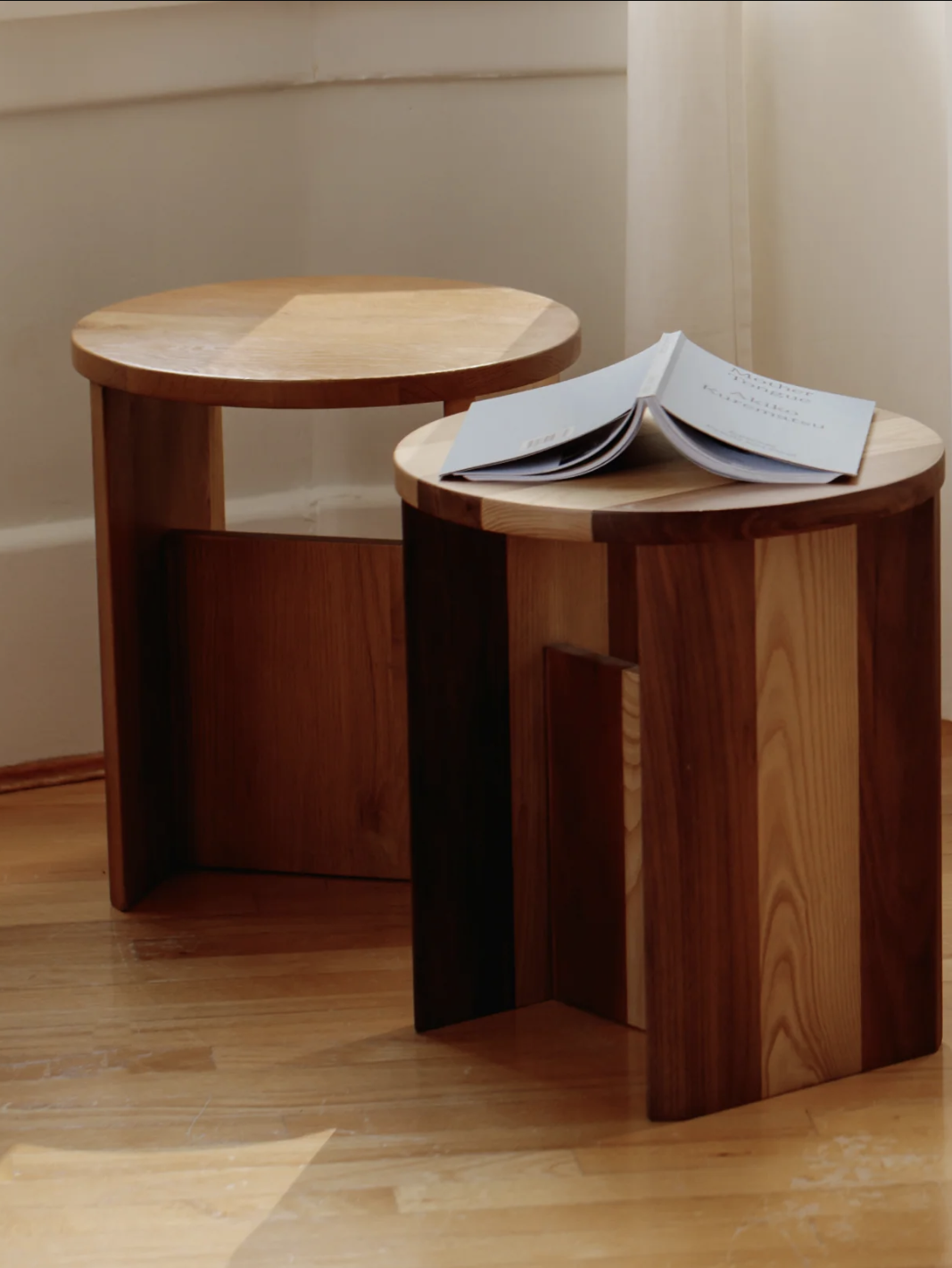 Two wooden stools in a corner of a sunlit room. An open book rests on one stool, with soft, sheer curtains in the background.
