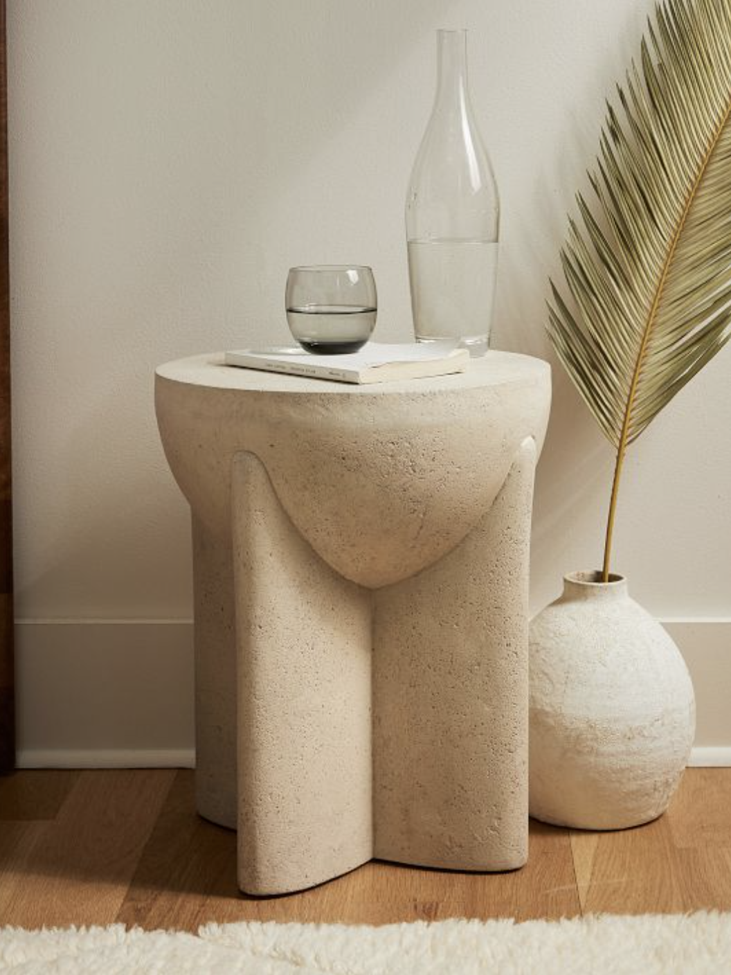 A small beige stone table holds a glass of water and a carafe. A white vase with a dried palm leaf is on the floor next to it, set against a neutral wall and a wooden floor.