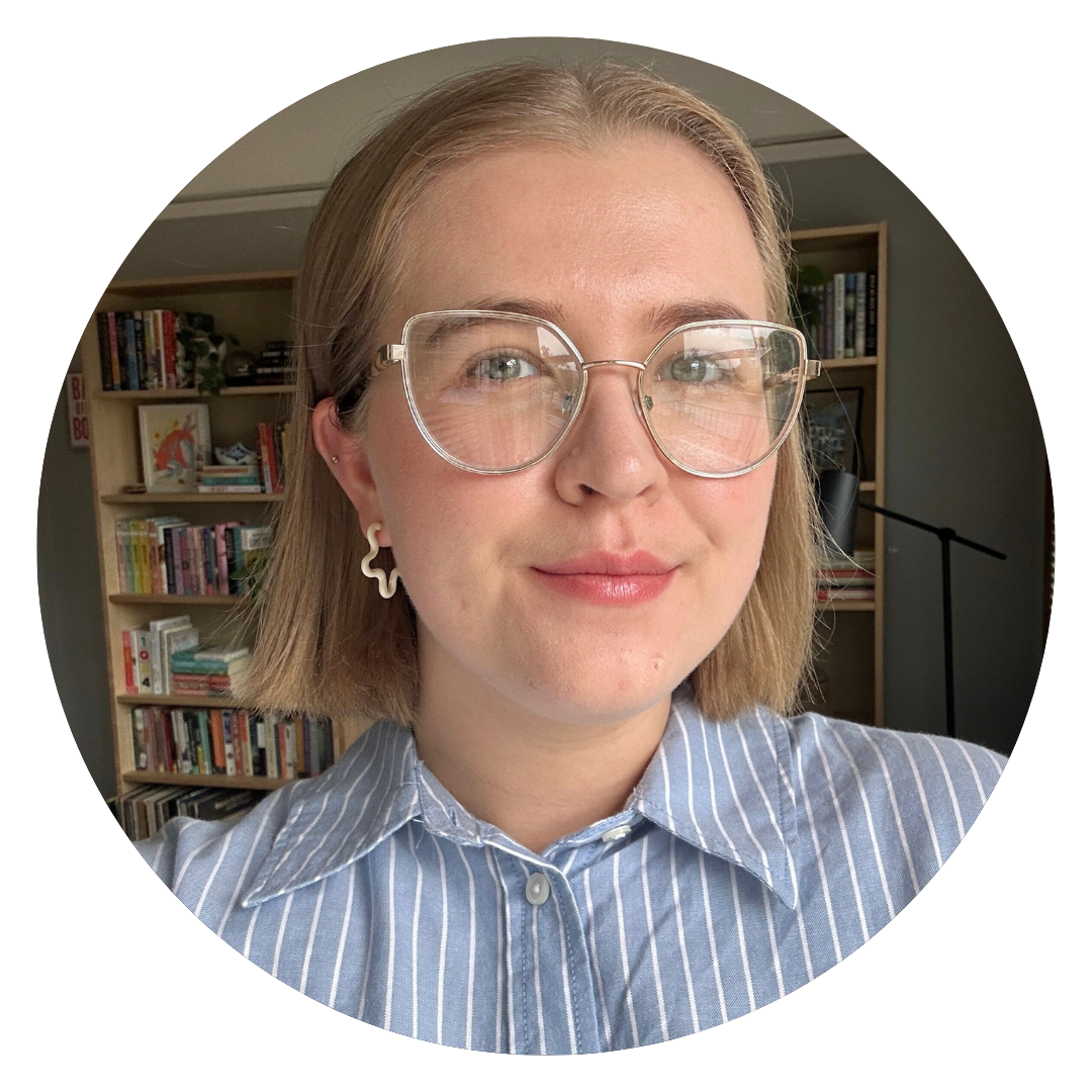Person with short blonde hair and clear glasses, wearing a blue striped shirt, stands indoors in front of bookshelves and a lamp.