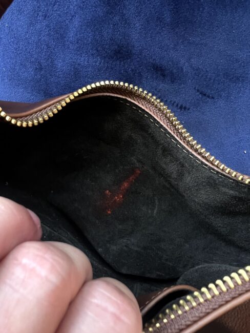 A hand opens a brown zippered pouch, revealing a dark interior with a small red mark or stain on the lining against a blue background—a detail often mentioned in a Cuyana review.