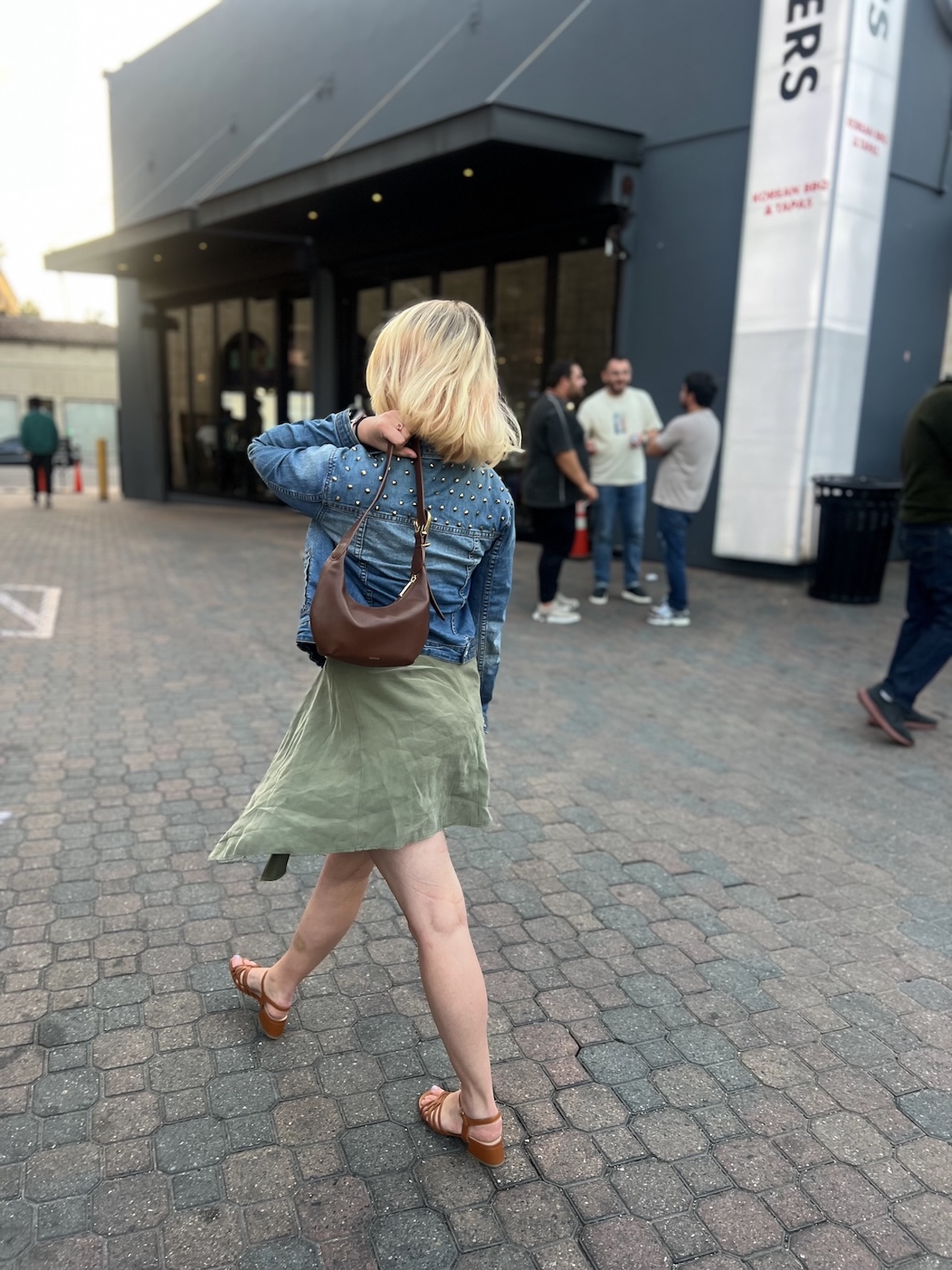 A woman with blonde hair, wearing a denim jacket and green skirt, walks on a paved path outside a building, carrying a brown purse over her shoulder—perfect for a Cuyana review. Several people stand in the background.