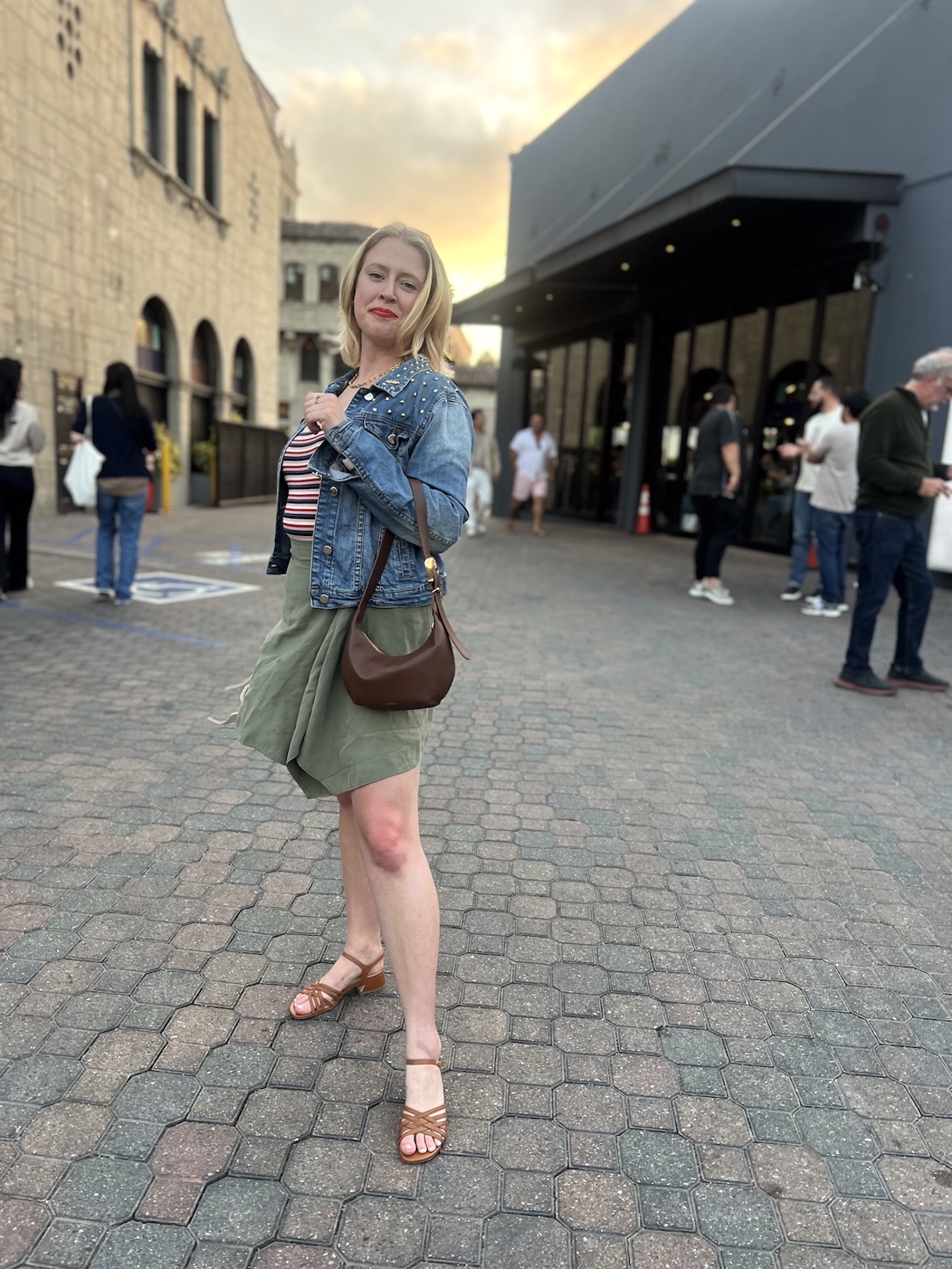 A woman stands on a paved street wearing a denim jacket, striped top, olive skirt, and sandals, holding a brown shoulder bag—perfect for any cuyana review. People and buildings appear in the background.