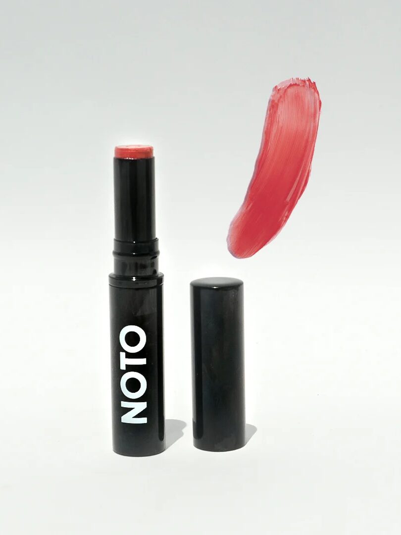 A black lipstick tube labeled "NOTO" stands upright with its cap beside it. A swatch of red lipstick is shown above.