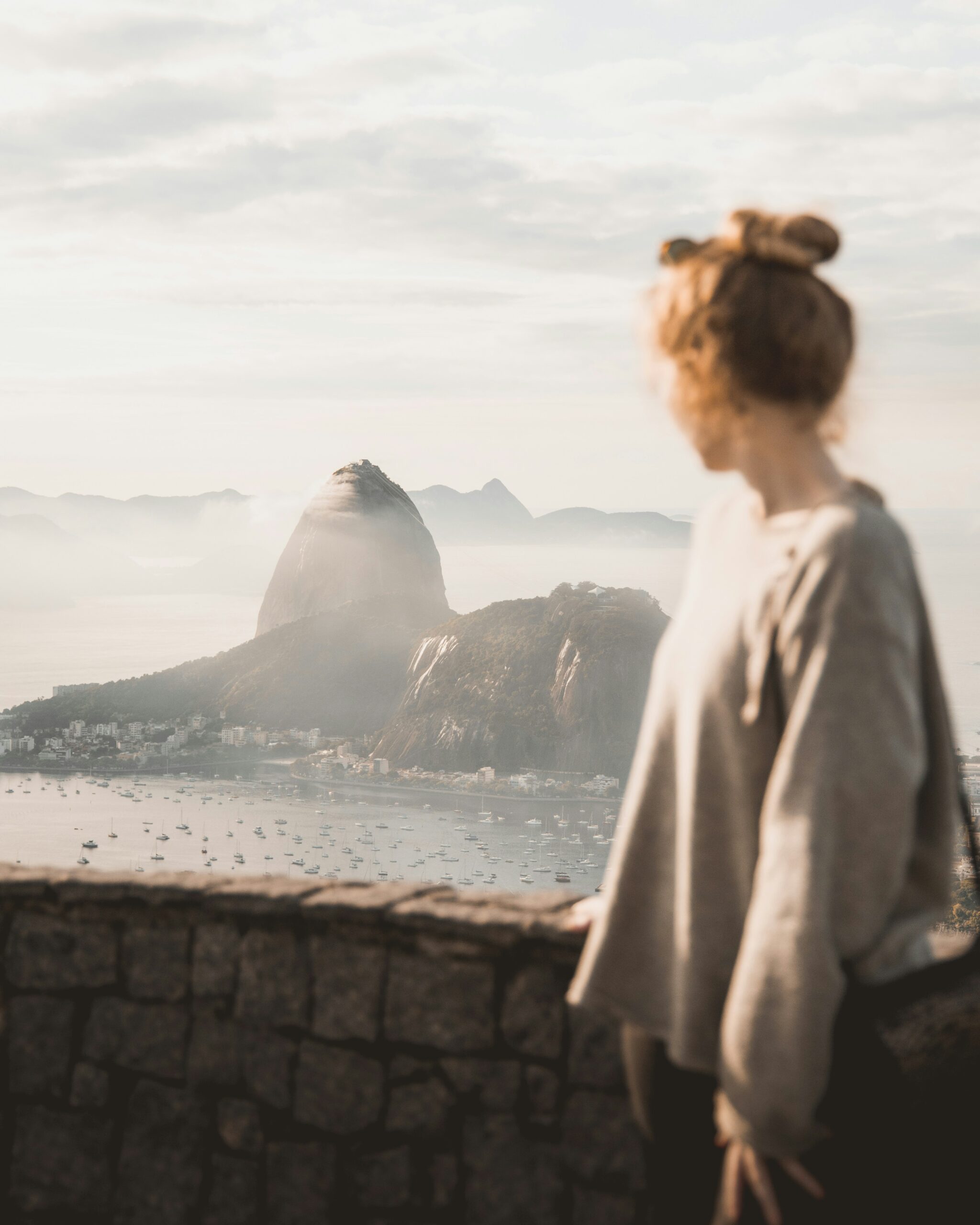 A person with light brown hair in a bun stands by a stone wall, overlooking Sugarloaf Mountain and the bay in Rio de Janeiro, Brazil, on a hazy day.