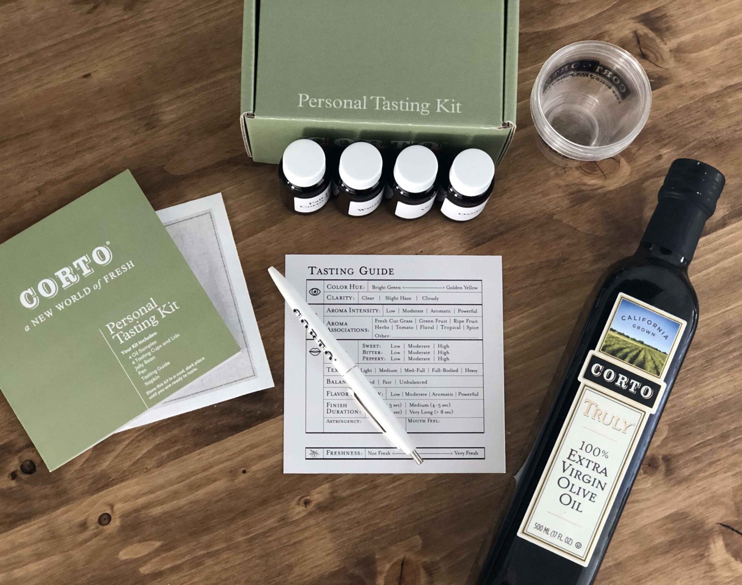 Personal tasting kit with olive oil bottle, four small sample bottles, tasting guide, instruction booklet, pen, and plastic cup on wooden table.