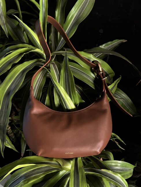 A brown leather Cuyana handbag with a shoulder strap is displayed hanging on green tropical leaves against a dark background—perfect for your next Cuyana review.