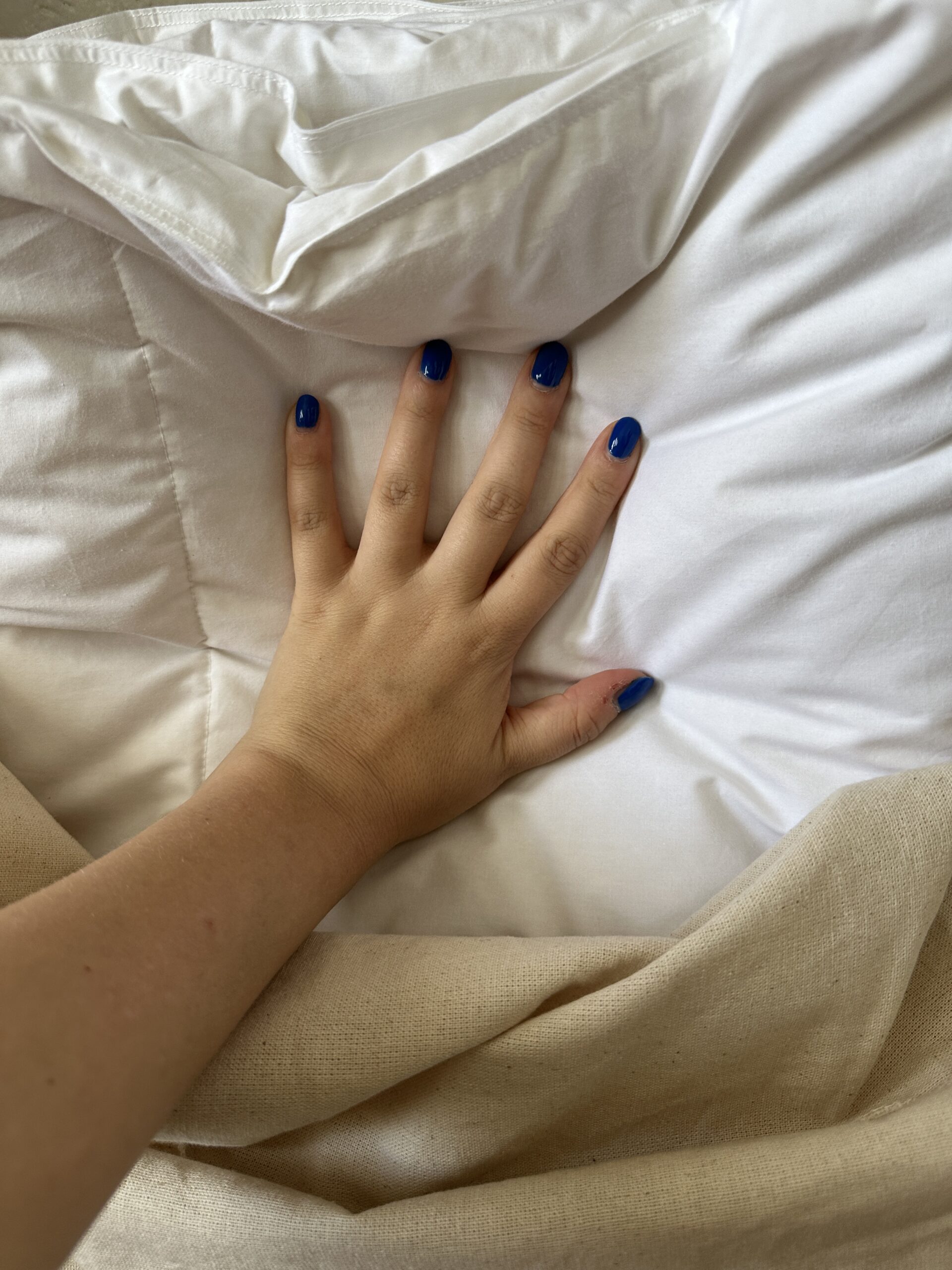 A hand with blue nail polish rests on a white quilted blanket beside a beige fabric edge.