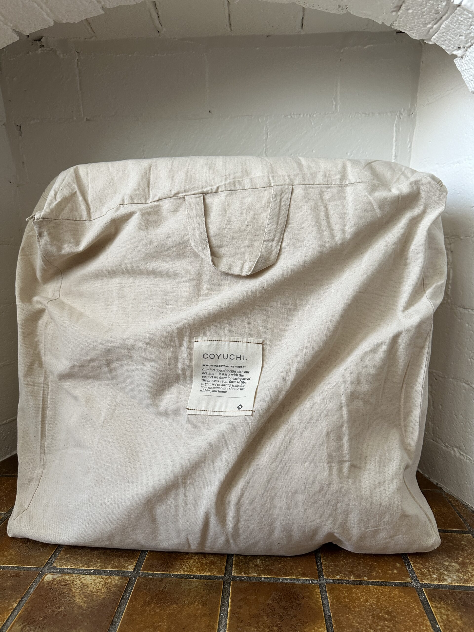A beige fabric bag with a handle and a label stands against a white tiled wall on a brown tiled floor.