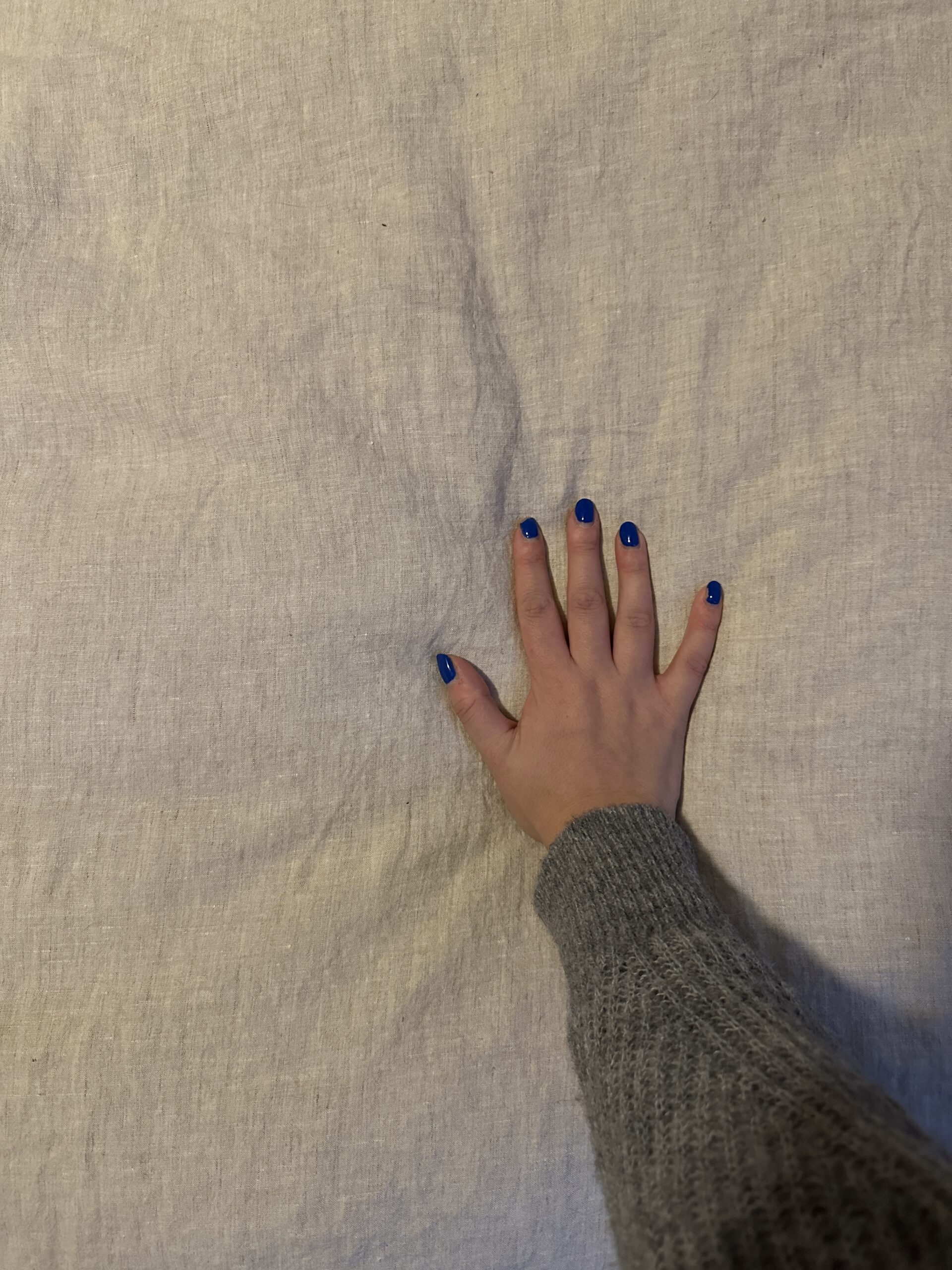 A hand with blue nail polish and a gray sweater sleeve rests on a light gray fabric surface.