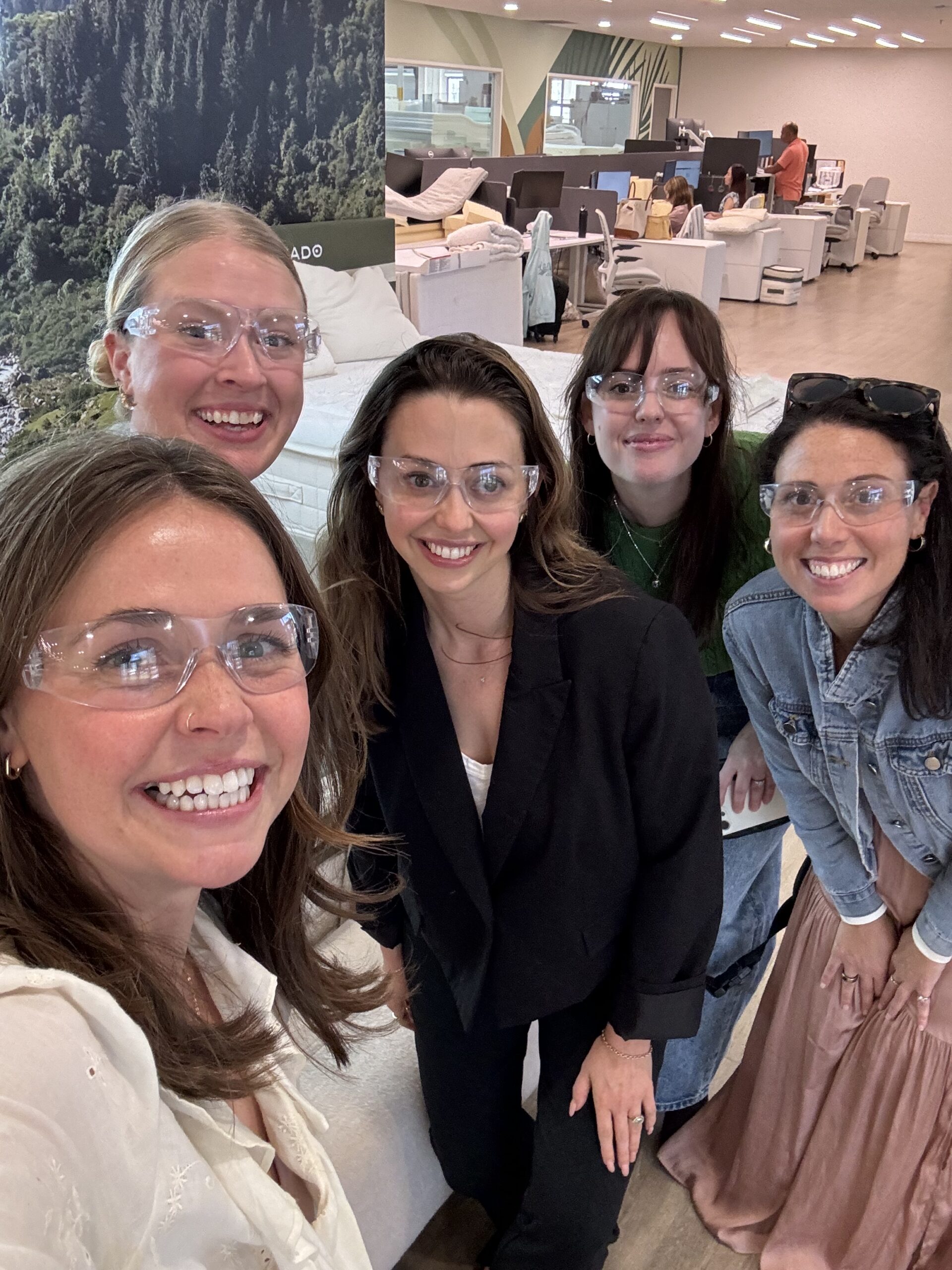 Five people wearing safety glasses smile at the camera inside a modern office space with a forest mural in the background.