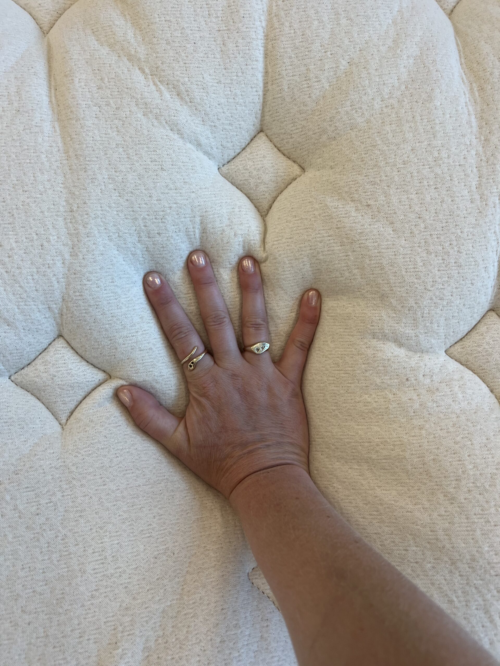 A hand with rings is pressing down on a quilted, cream-colored mattress, creating an indentation.