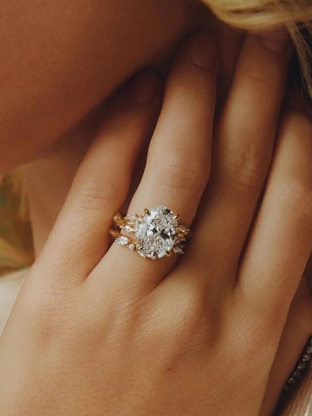 A person wears a gold ring with a large round diamond, accompanied by a matching band, against a light background.