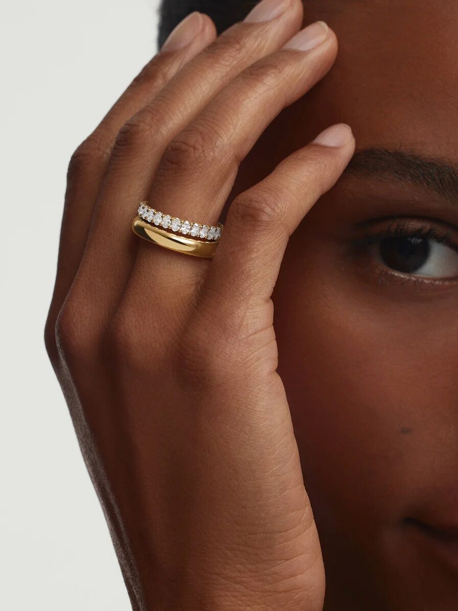 A person gently touches their face with their hand, showcasing a gold ring stacked with a diamond-studded band on their finger, against a plain background.