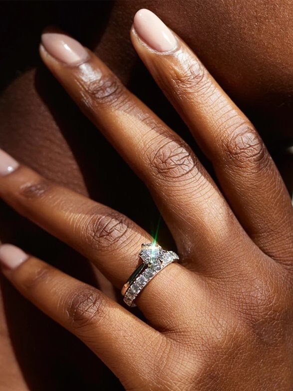 A close-up of a hand wearing a diamond engagement ring set against a blurred background. Text at the bottom reads "Shown with 1.50ct.