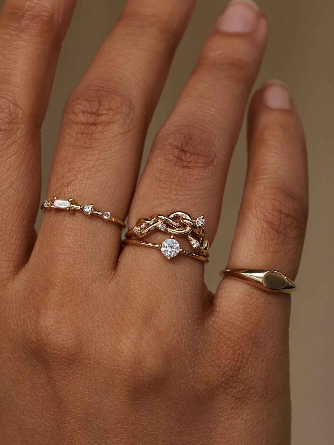 A hand wearing four gold rings, including a braided band with a central diamond, a slim band with small stones, and a plain band with an engraved pattern.