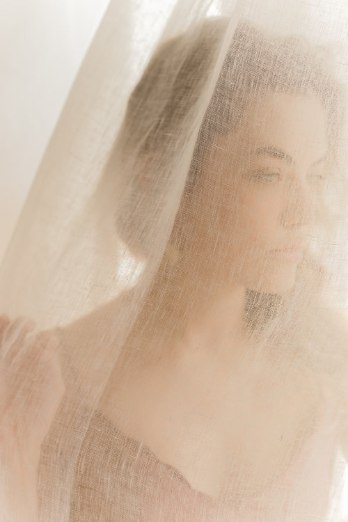 A woman standing behind a sheer curtain, softly focused, looking to the side.