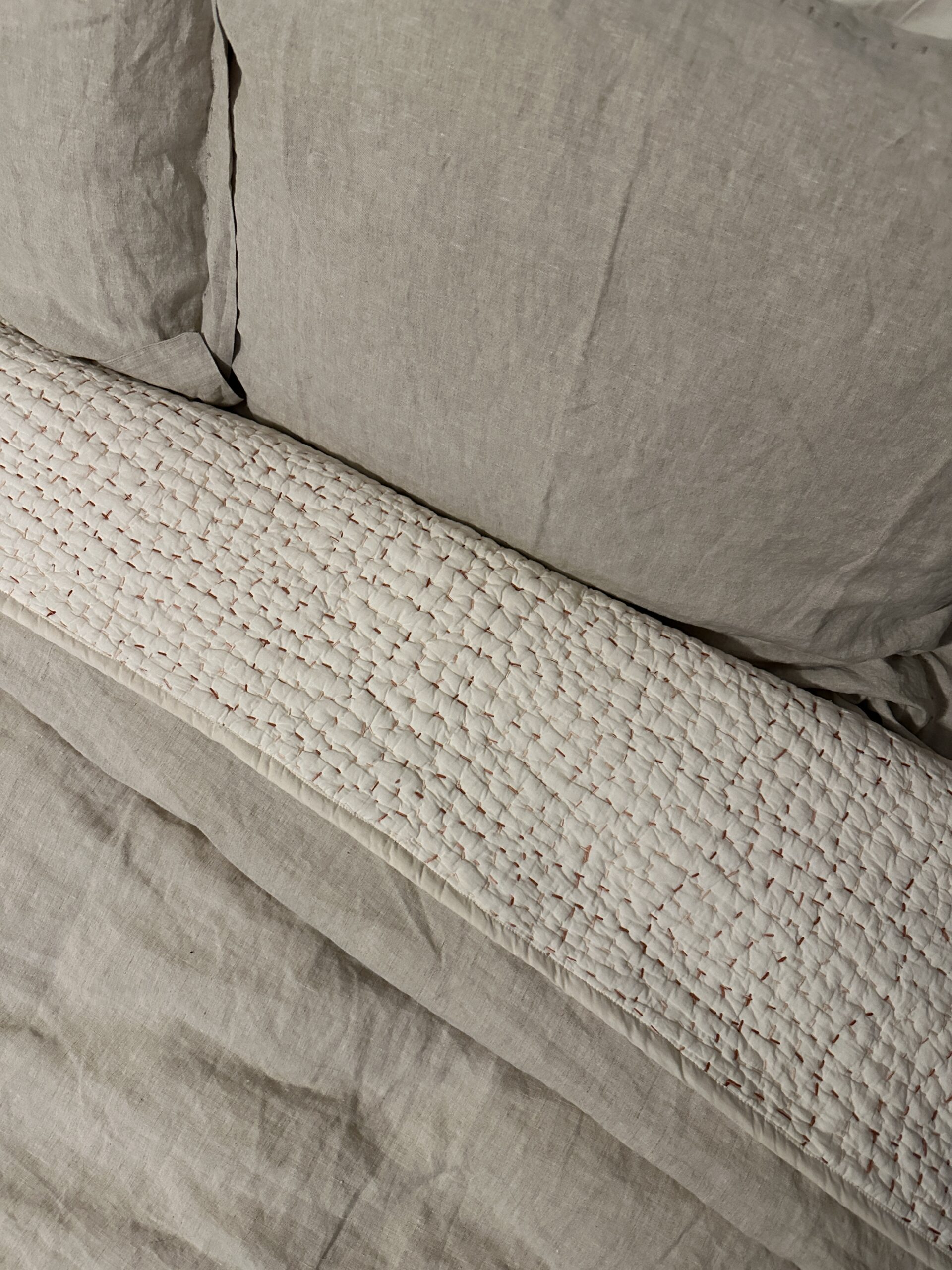 A neatly made bed with beige linen sheets, a matching pillow, and a textured white quilted blanket folded beneath the pillow.