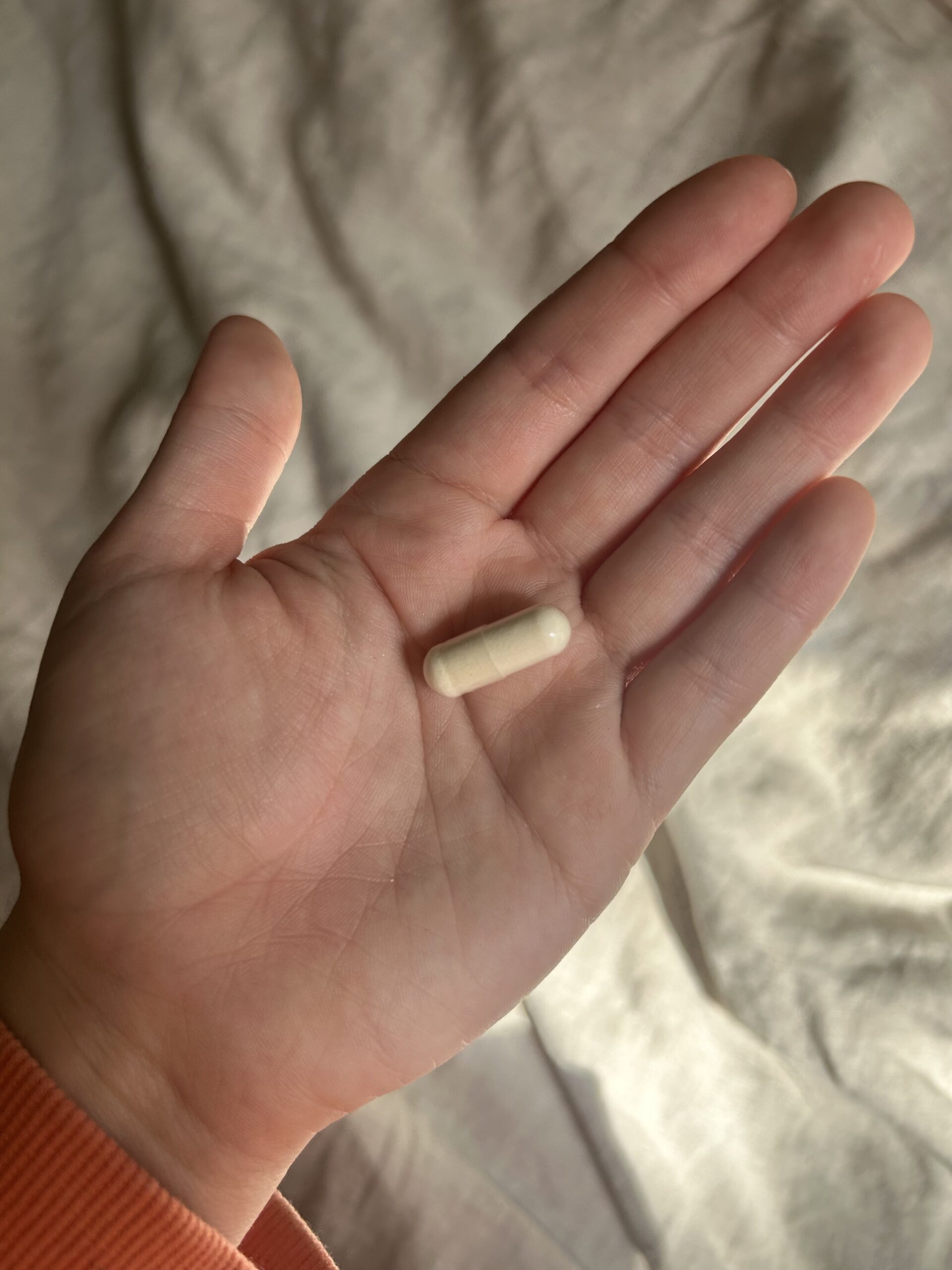 A hand holding a single white capsule-shaped pill against a light-colored background.