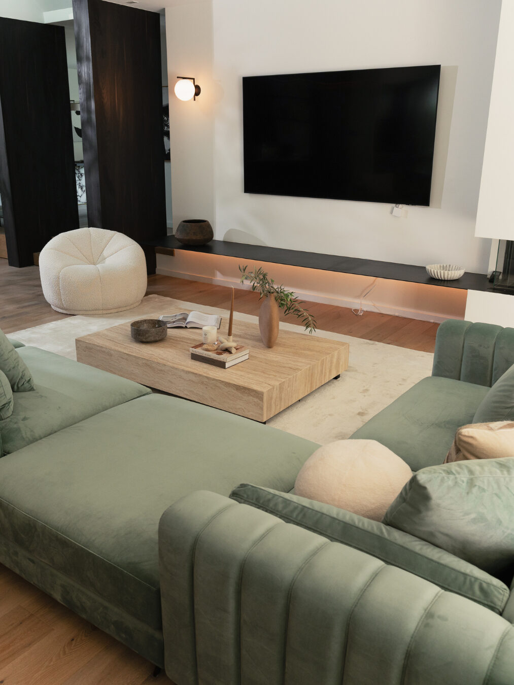 Modern living room with a large TV on the wall, green sectional sofa, round white chair, and a wooden coffee table with decor items on a light-colored rug.