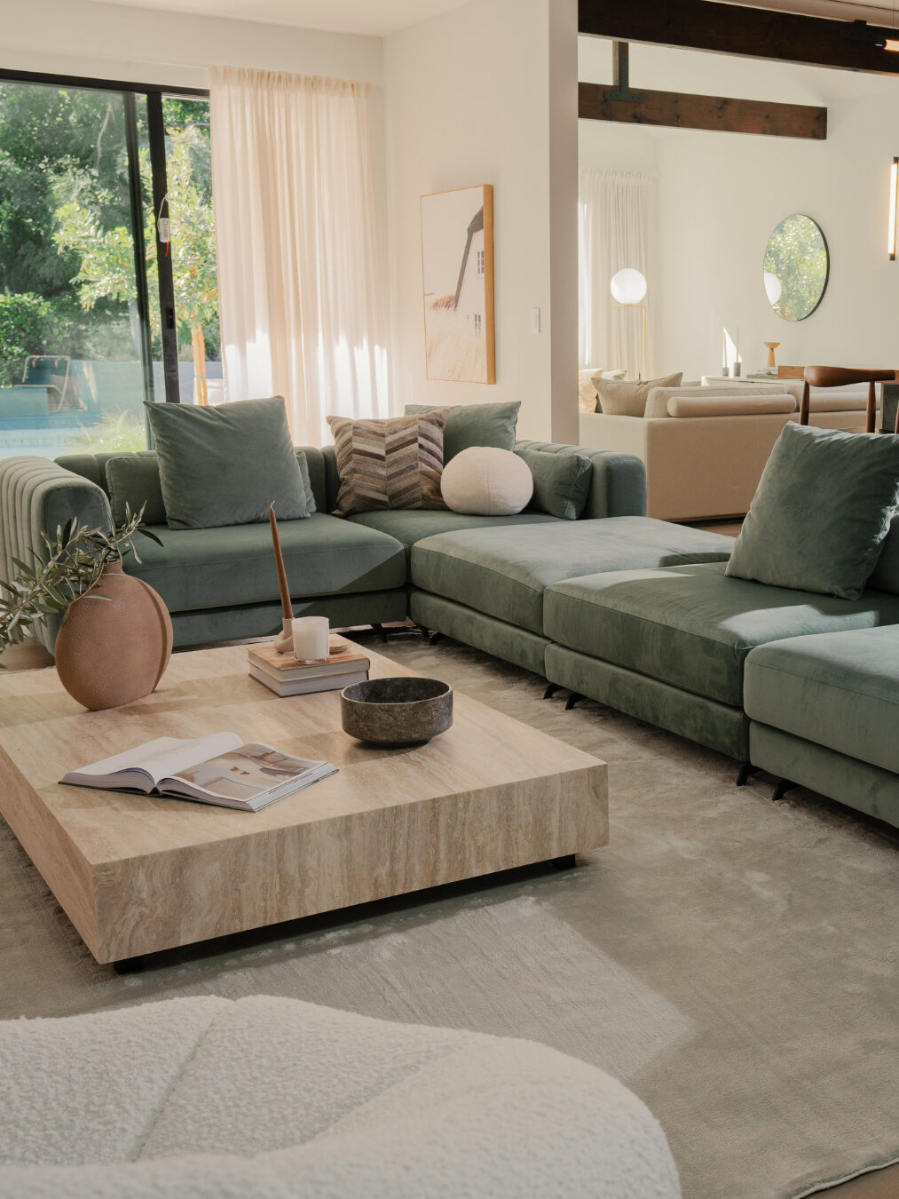 Modern living room with a large green sectional sofa, a low wooden coffee table, and a view of a backyard pool through glass doors. Neutral decor and natural light fill the space.