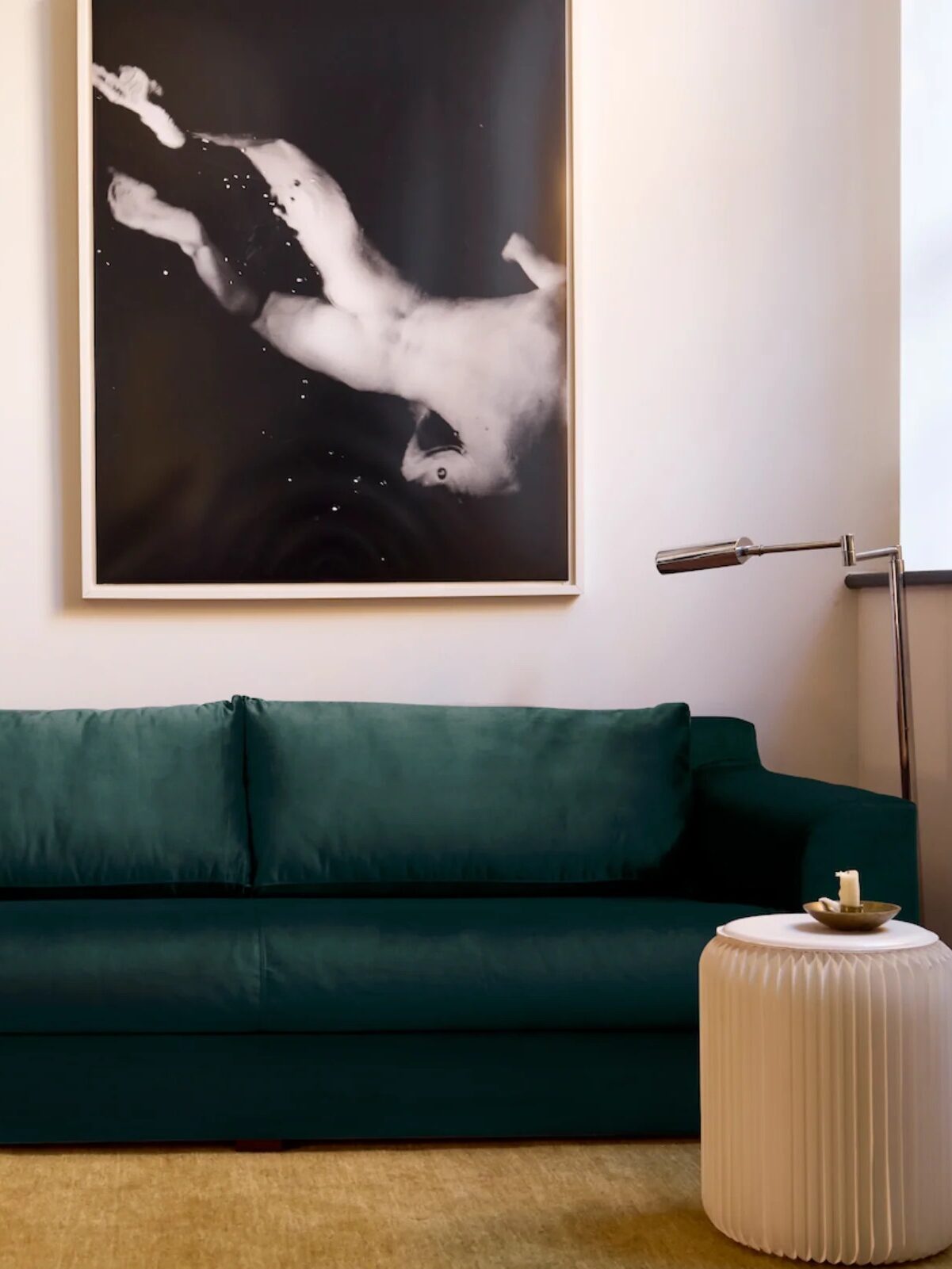 A modern living room with a dark green sofa, mustard pillow, abstract wall art, and a side table with a lamp. A folded blanket hangs on a stand beside a small cart.