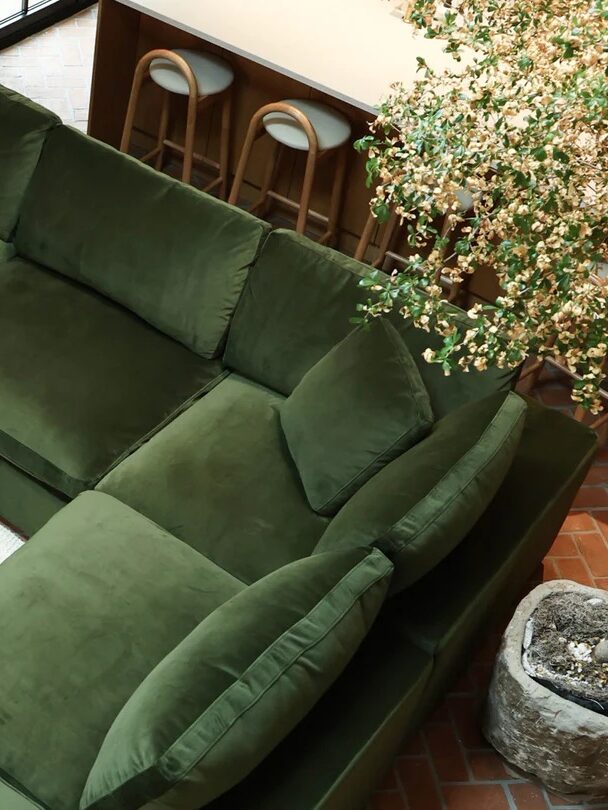 A green sectional sofa in a living room with a concrete coffee table topped with books and a decorative item. Bar stools are visible in the background near a counter.