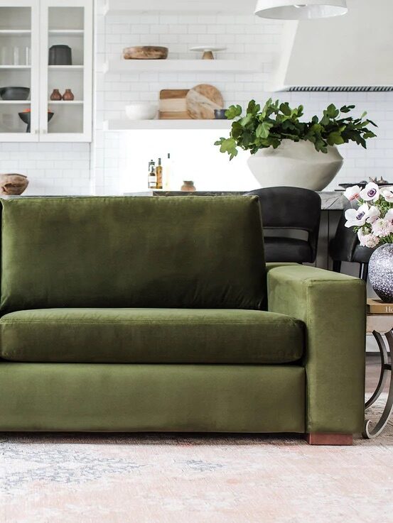 A green velvet sofa with cushions and a throw blanket in a modern living room. In the background are glass cabinets, plants, and decorative items.