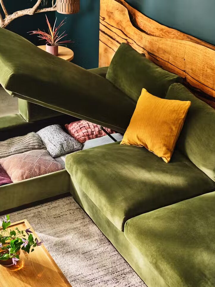 Green velvet sectional sofa with open storage compartment containing colorful pillows and blankets. Wooden backrest detail, coffee table with plant, and decorative items in the background.