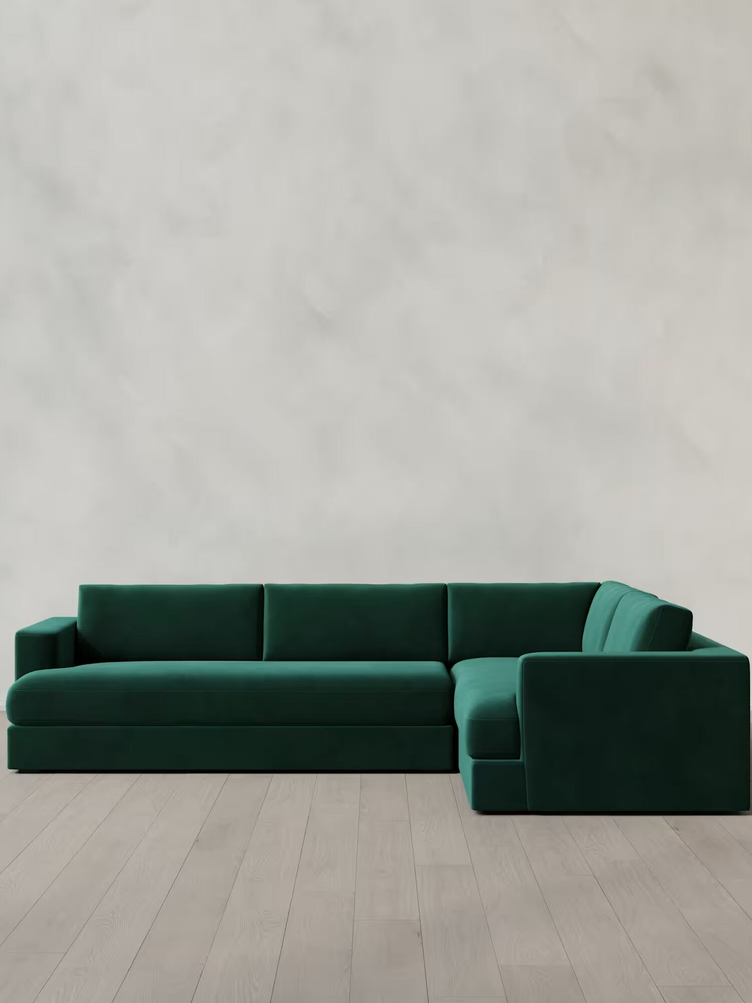 A green velvet L-shaped sectional sofa on a light wood floor against a plain off-white wall.