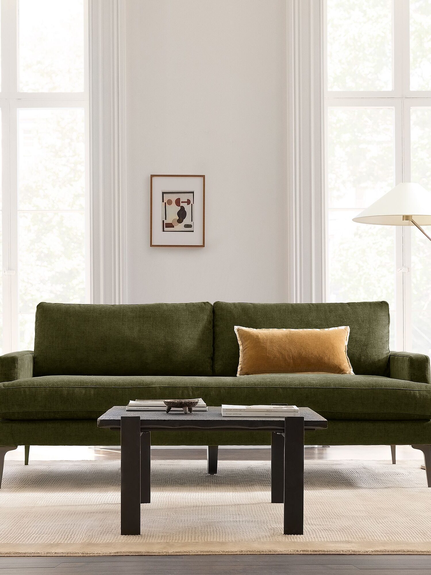A green sofa with a yellow cushion, a black coffee table, a modern floor lamp, and framed art are set against a light-filled room with large windows.
