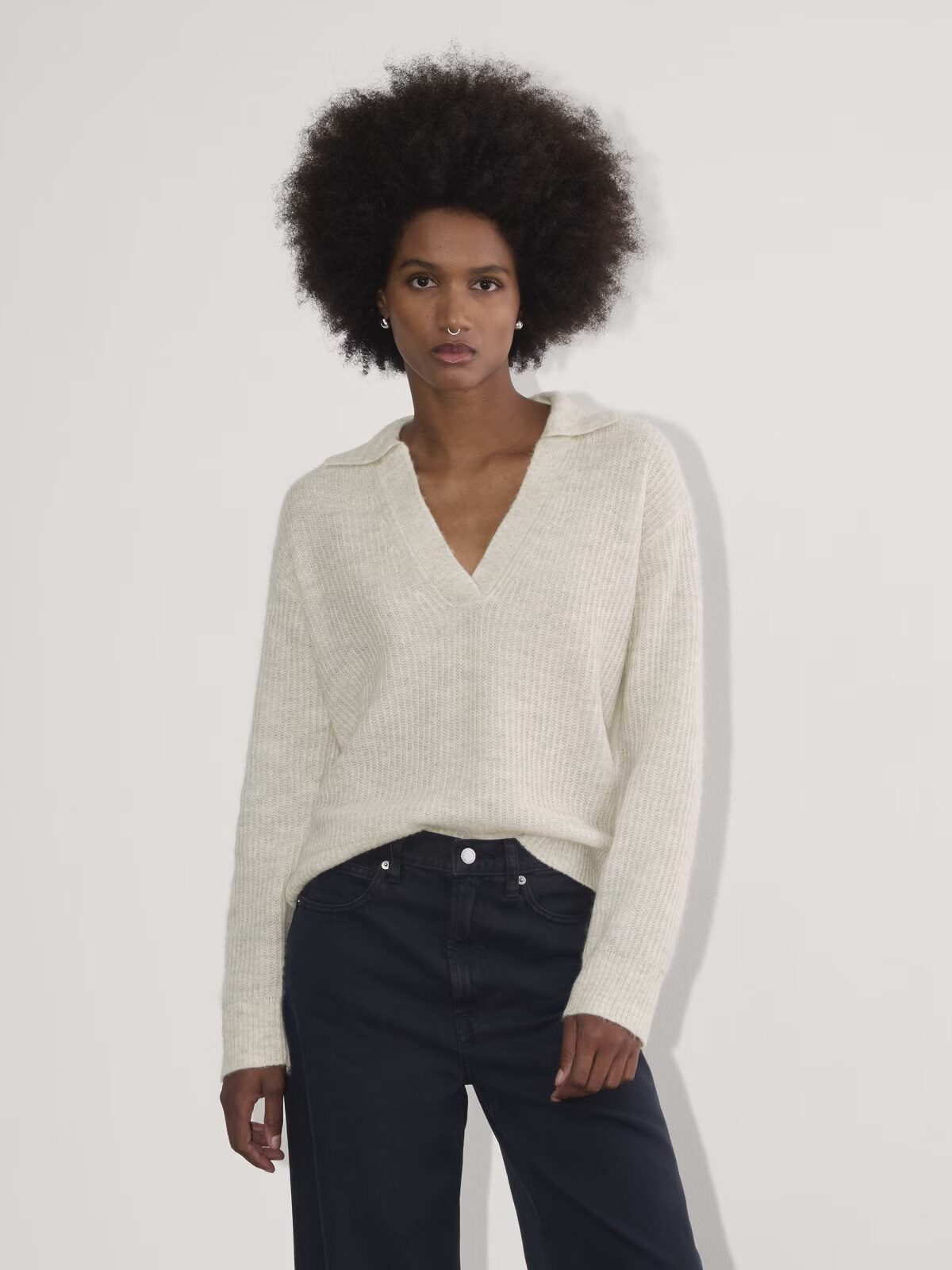 Person with an afro hairstyle wearing a cream-colored sweater and black pants, standing against a plain white background.