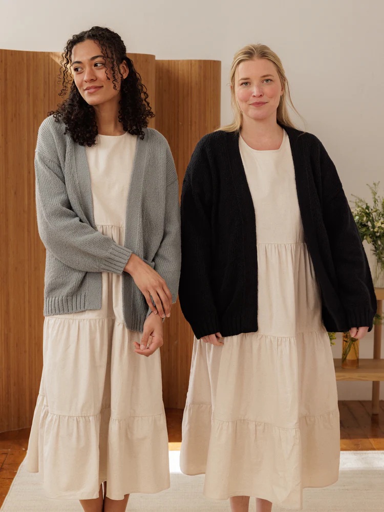 Two people stand indoors wearing cream dresses with knitted cardigans, one gray and one black, against a simple background with a wooden screen and flowers.