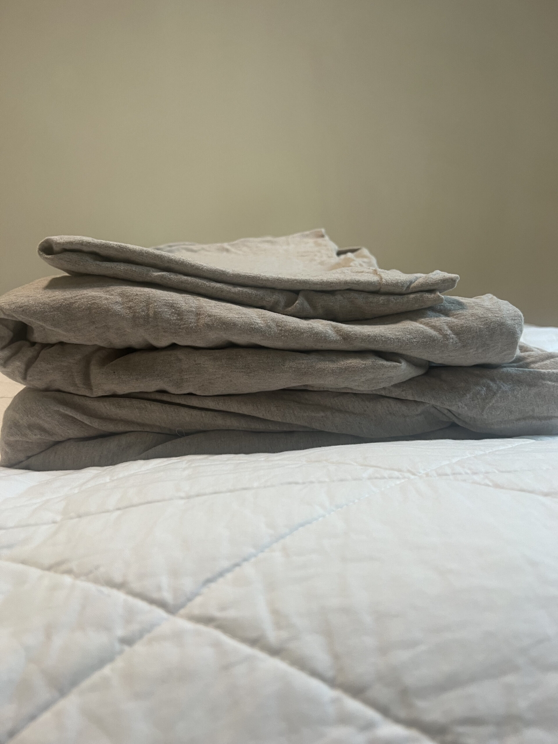 A neatly folded stack of gray sheets rests on a white quilted bedspread against a plain light-colored wall.