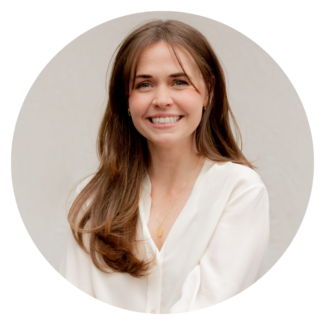 Woman with long brown hair, wearing a white blouse and gold necklace, smiling at the camera against a neutral background.