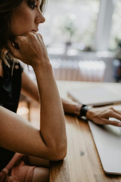 Person seated at a wooden table, wearing a watch, looking at a laptop screen with hand on chin in a thoughtful pose.
