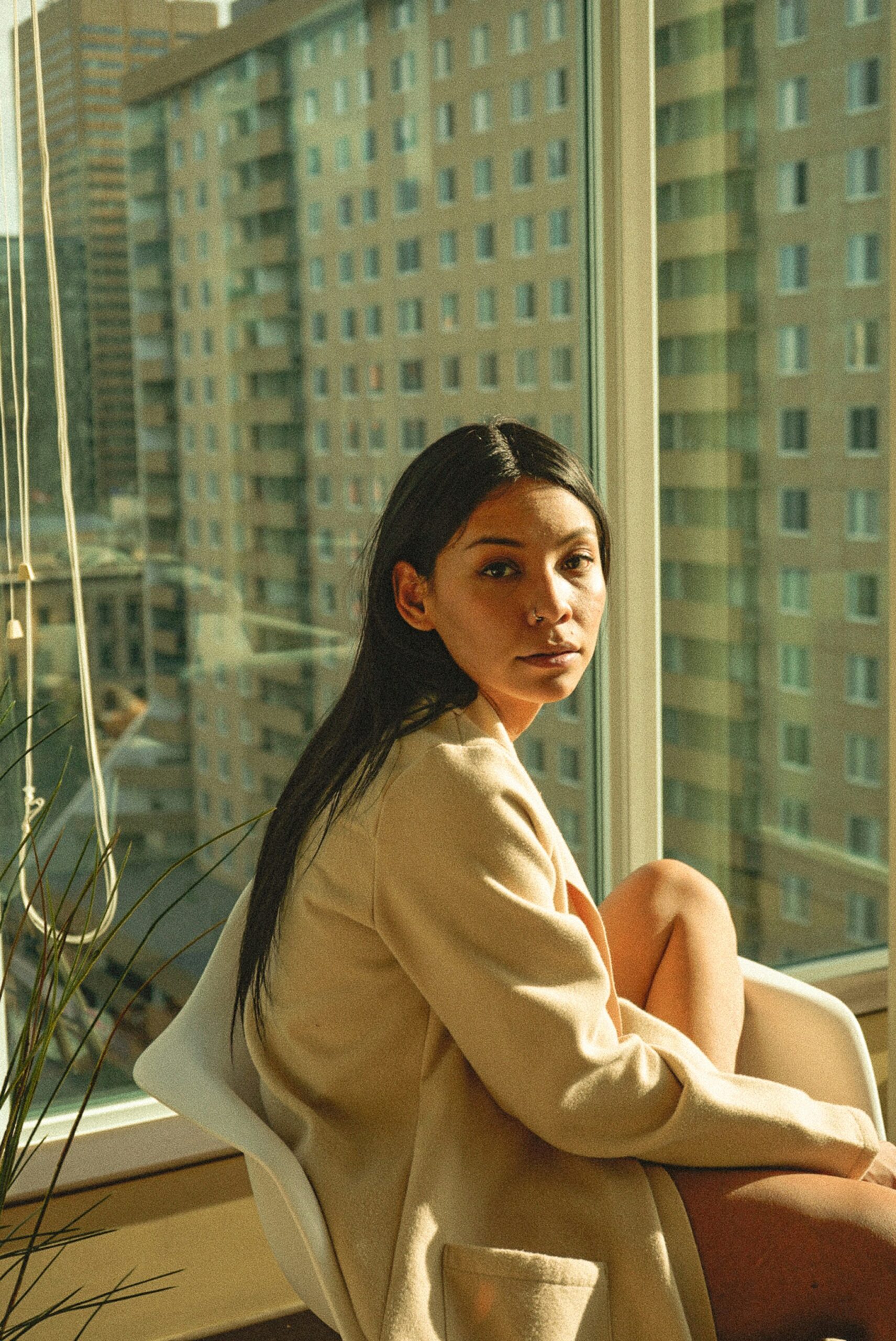 Person sitting on a chair by a window, wearing a light-colored coat. High-rise buildings are visible outside.