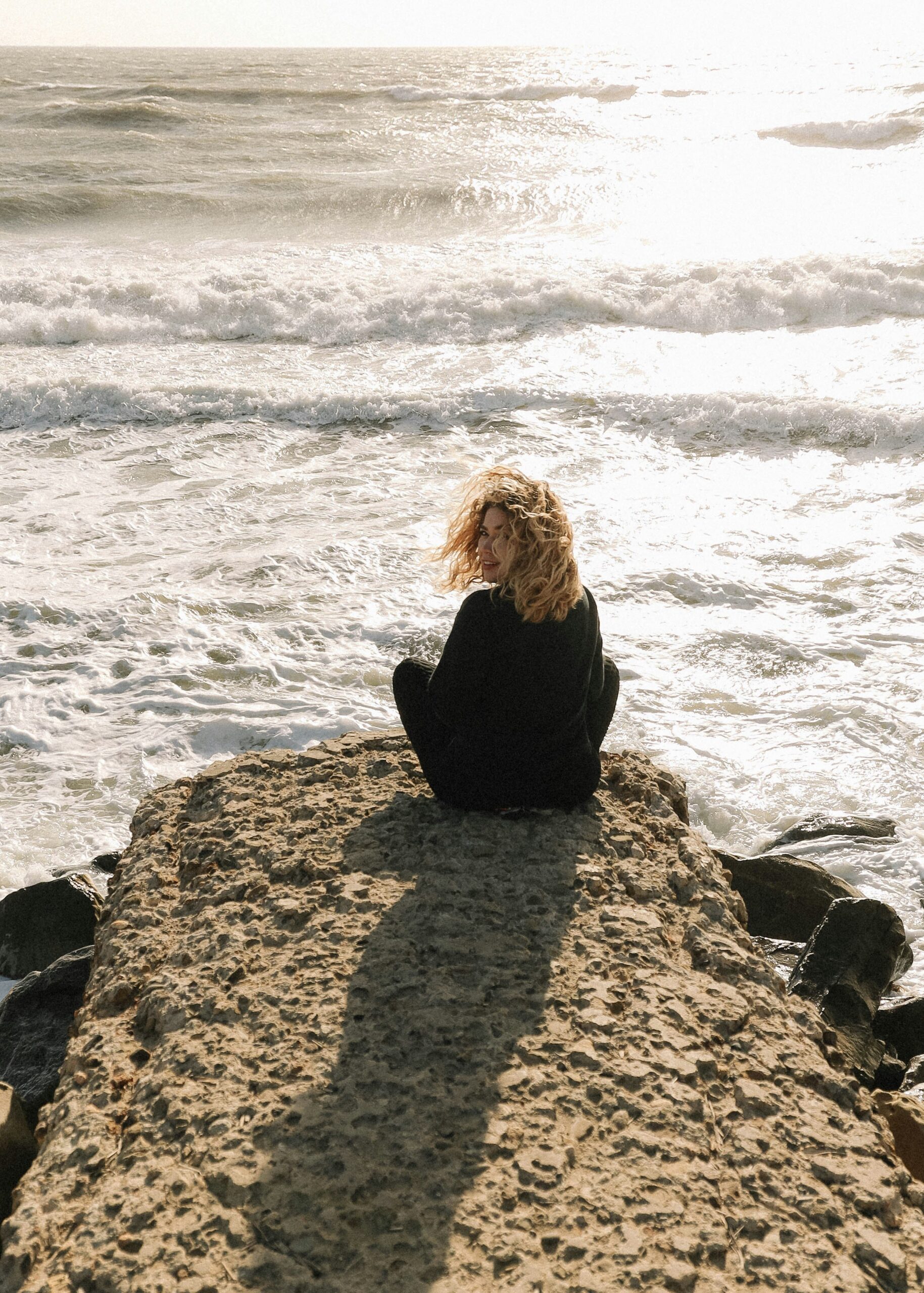 Person with curly hair sits on a rocky outcrop, facing a sunlit ocean with waves breaking, under a cloudy sky.