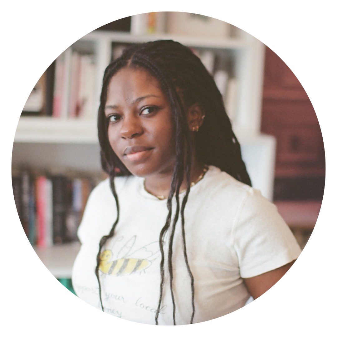 A person with long, braided hair wearing a white t-shirt stands indoors in front of a blurred bookshelf.