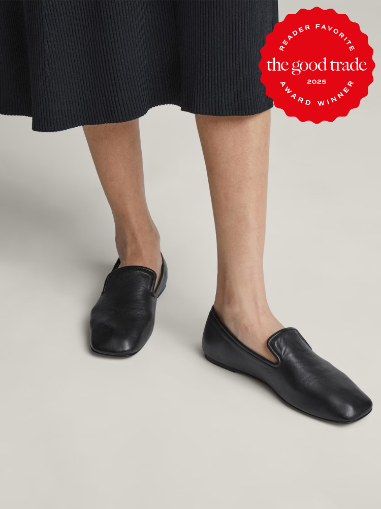 Woman wearing black slip-on loafers and a black skirt. A red award badge for "The Good Trade" is in the top right corner.