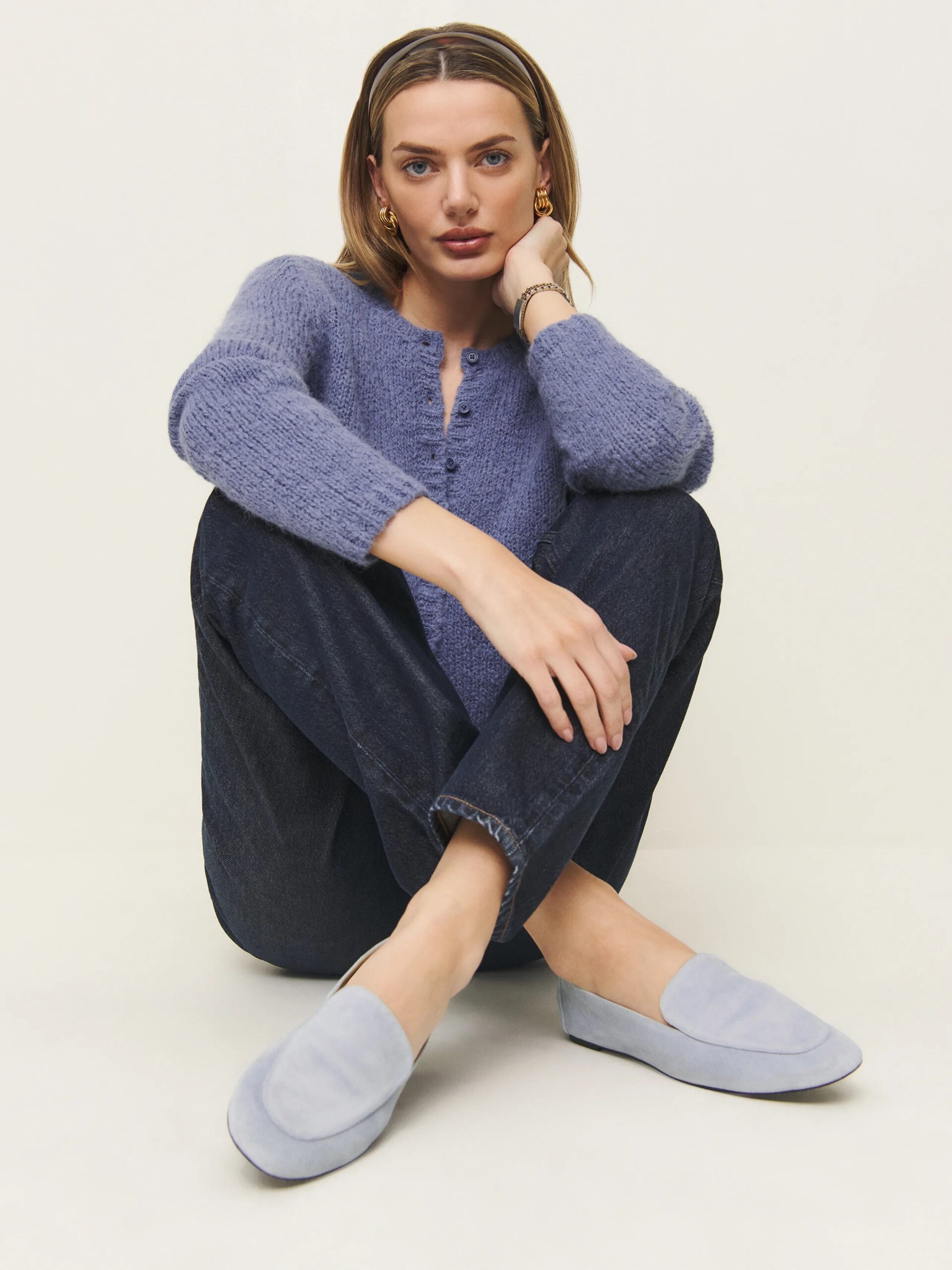 A person sits cross-legged on the floor, wearing a blue sweater, dark jeans, and light blue slip-on shoes, against a plain backdrop.
