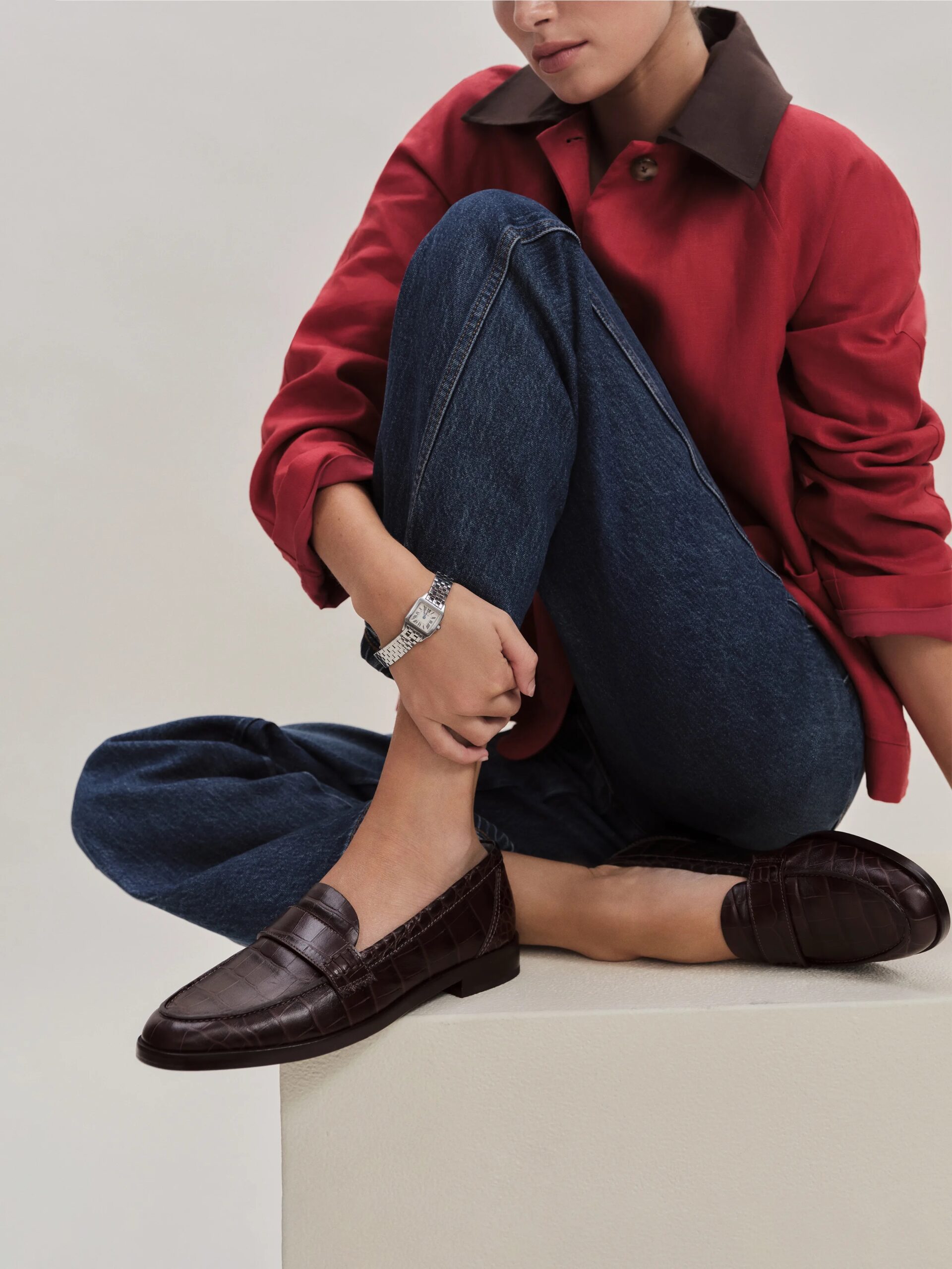 Person sitting on a white block, wearing a red jacket, blue jeans, and brown loafers.
