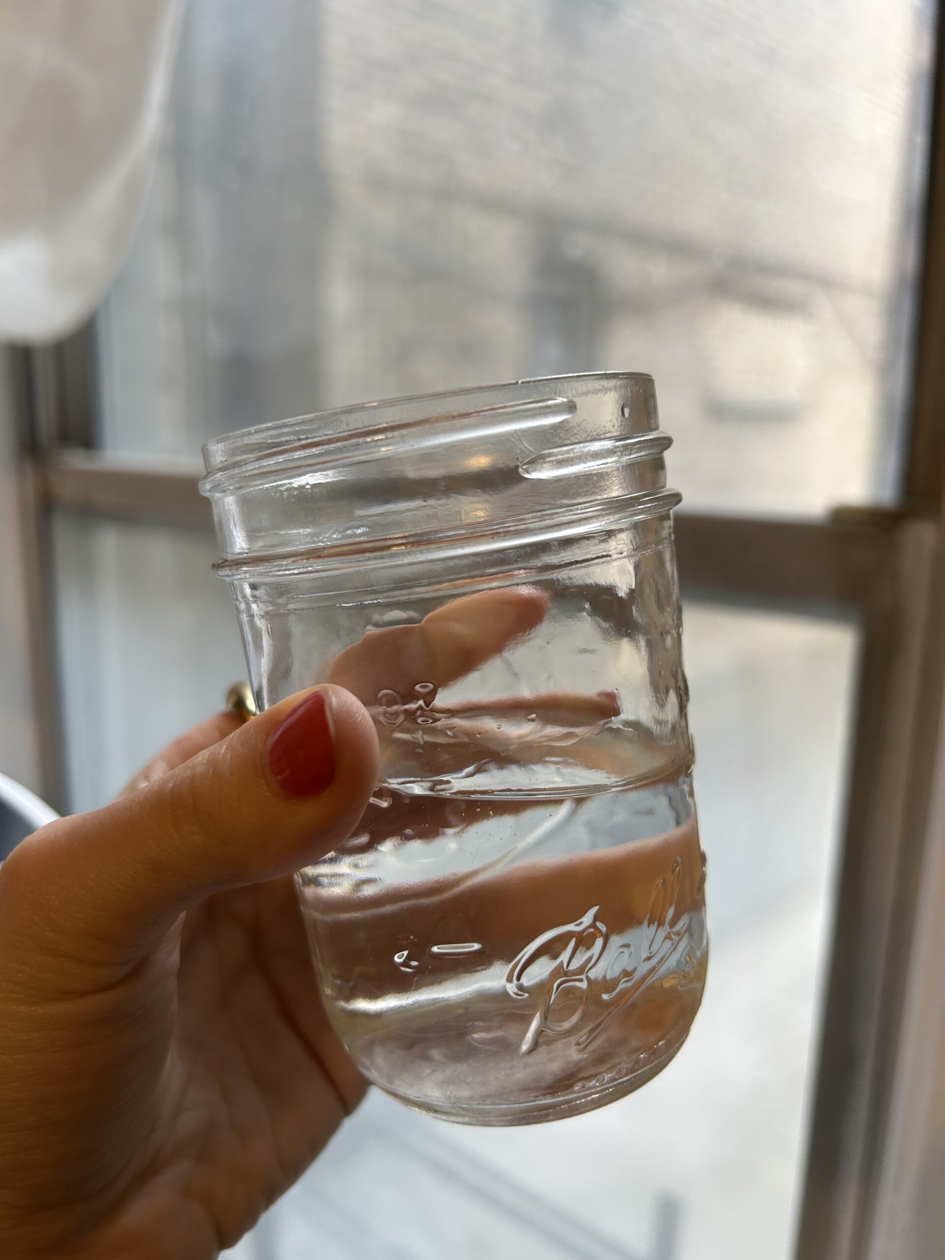 A hand with red nail polish holds a mason jar partially filled with water near a window.