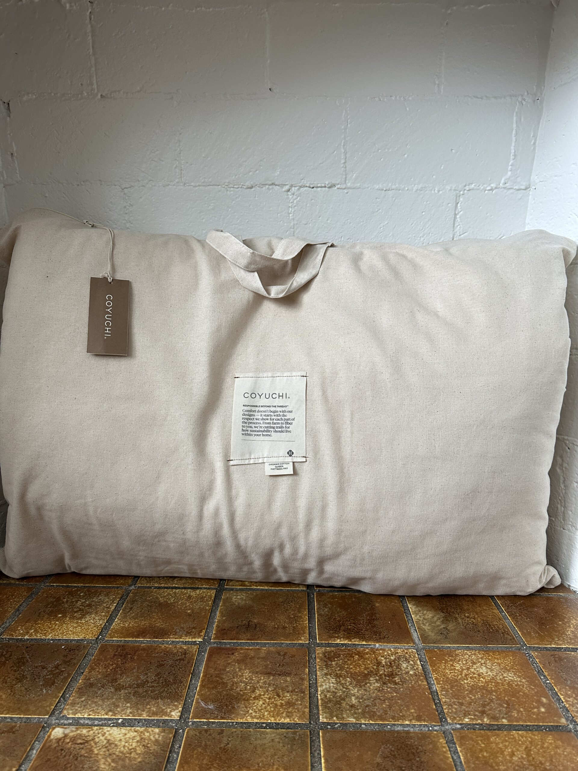 A large, cream-colored cloth bag with handles is placed on a tiled floor against a white wall. It has a label on the front and a tag hanging from the handle.