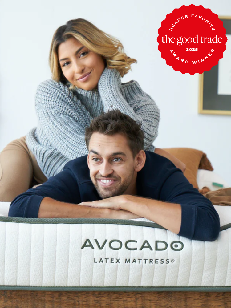 A man and woman smiling, leaning on an Avocado latex mattress. The image features a "The Good Trade 2023 Reader Favorite Award Winner" badge in the top right corner.