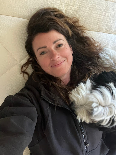 A person with long, wavy hair is lying on a bed, smiling. They are wearing a dark jacket and resting beside a fluffy, black-and-white object.