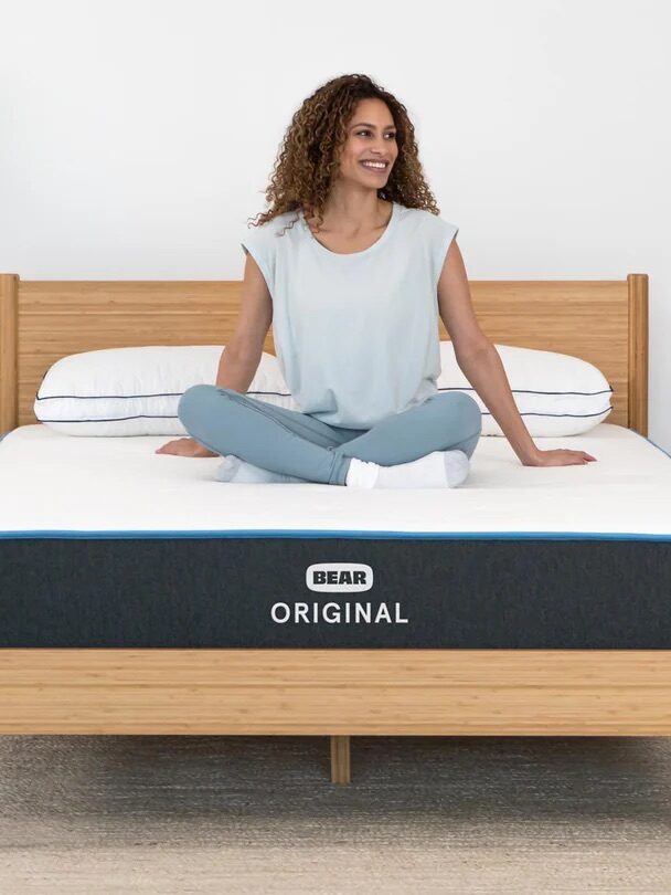A person sits cross-legged on a bed with a "Bear Original" mattress. The room features a basket, lamp, and chair with an American flag pillow.