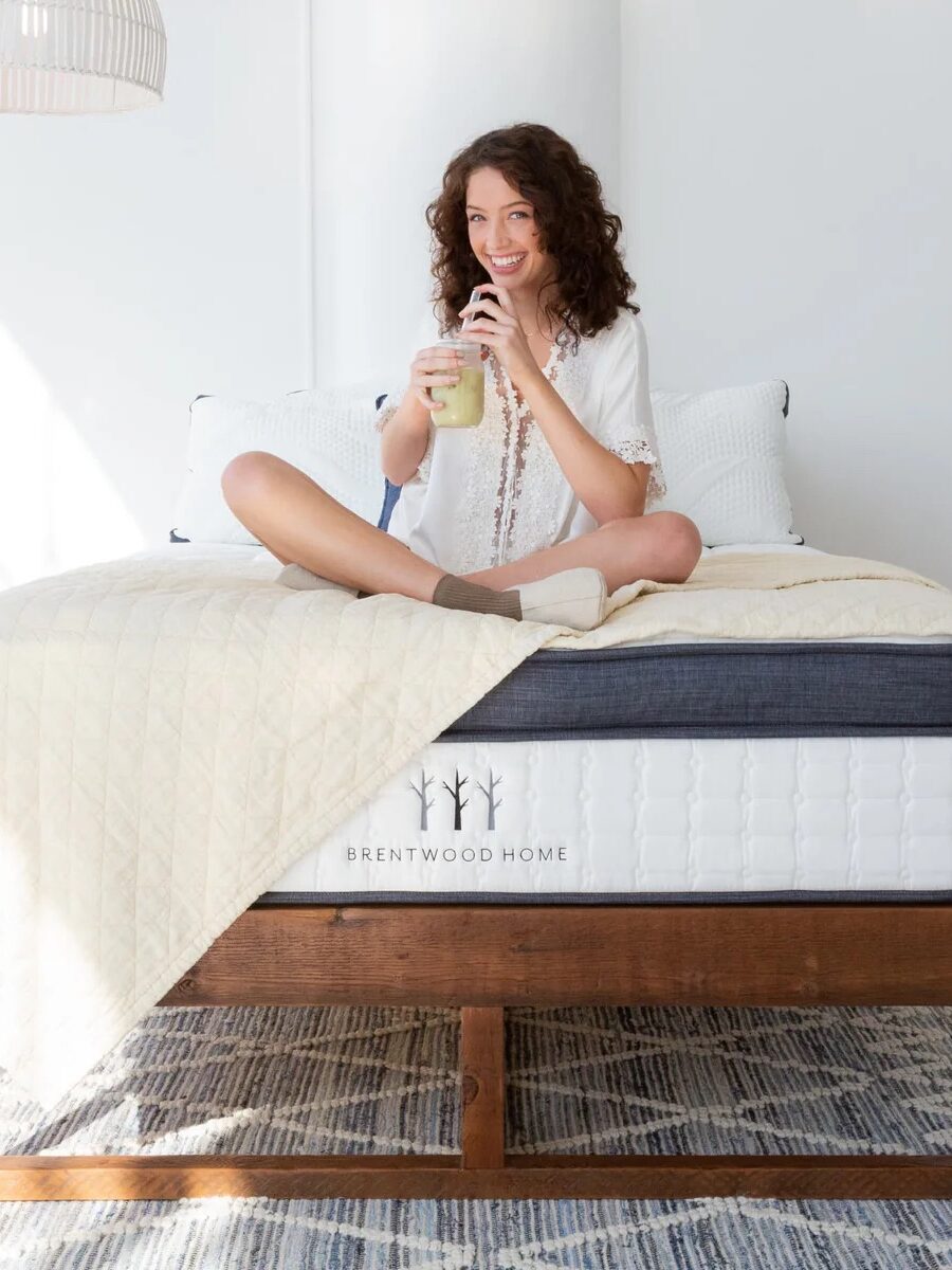 Person sitting cross-legged on a bed, drinking from a straw. The bed is branded Brentwood Home with cream bedding and a geometric rug underneath.