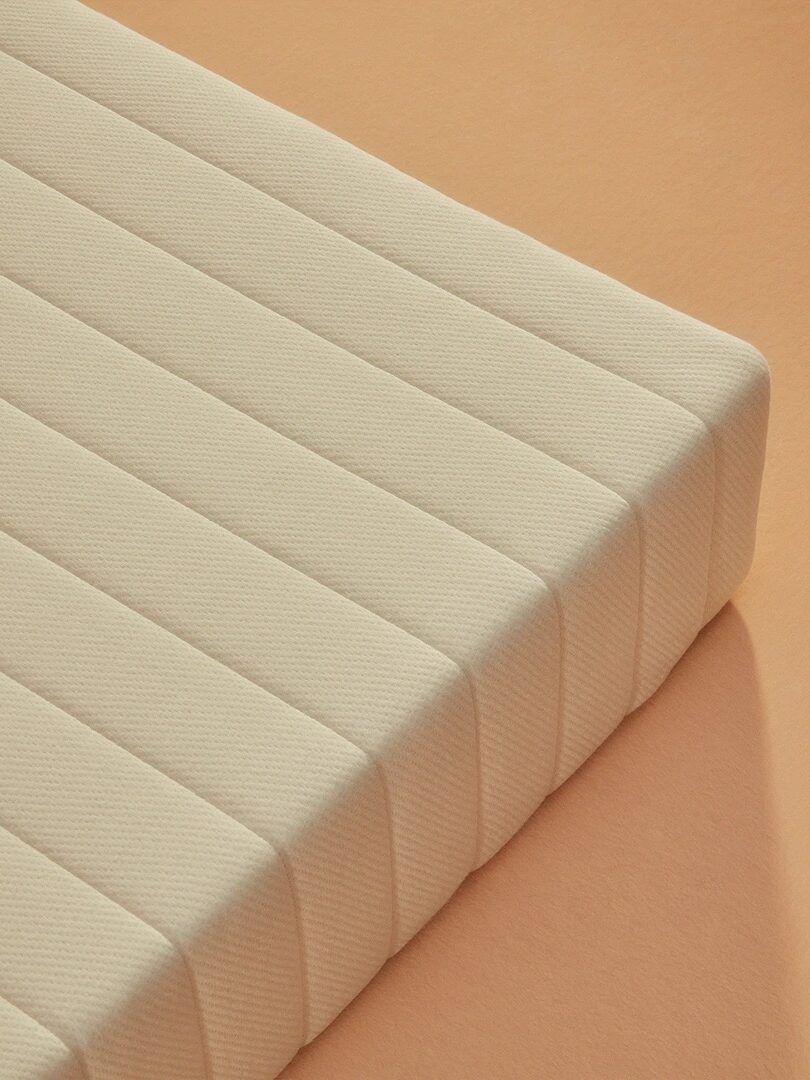 A beige mattress with striped quilting sits on an orange surface, viewed from an angled side perspective.